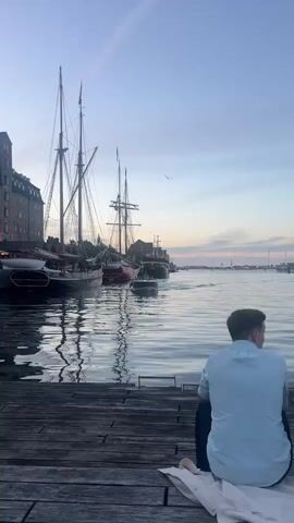 The media file shows a waterfront scene at dusk in Copenhagen, Denmark. The primary subject is a man sitting on a wooden pier, facing away from the camera and looking out at the water. The water is calm with gentle ripples reflecting the sky and the docked boats.

Several boats are visible, including large sailing vessels with tall masts and a smaller motorboat moving through the water. In the background, a long, multi-story brick building lines the waterfront, with many windows illuminated from within, suggesting it is evening. The sky displays soft colors of twilight, with hints of blue, pink, and orange, indicating the sun has recently set.

Further down the pier, a group of people are gathered. One man is seen walking towards the camera carrying a plastic bag. A white tour boat is docked nearby. The atmosphere appears peaceful and relaxed, with people enjoying the waterfront views as the day transitions into night. The overall scene depicts a serene urban harbor setting at dusk.

There is some visible text on the side of a white boat in the foreground that reads "SPIRAL HOTEL".