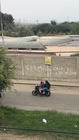The video shows an outdoor scene with a street and a brick wall with graffiti. On the street, there are several motorcycles and a car. One motorcycle has two people on it, while another motorcycle has one person. A white car is parked on the side of the street. The brick wall has some yellow signs and graffiti written on it. In the background, there are trees and buildings. The sky is cloudy.