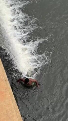 A person is swimming in dark water next to a concrete structure with water cascading over it. The water is agitated with white foam where it falls. The person is in the lower left quadrant of the frame, facing toward the cascading water. The overall scene appears to be a dam or a similar water control structure.