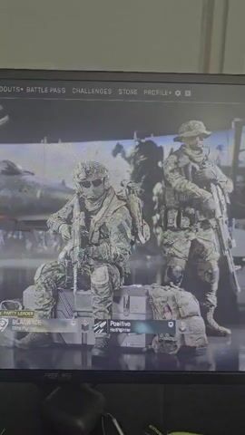 The video shows a computer screen displaying a video game menu. Two virtual characters, dressed in camouflage military uniforms, are standing on a platform in front of what appears to be a jet or airplane. The character on the left is seated, while the character on the right is standing. Text overlays on the screen indicate game options such as "LOADOUTS", "BATTLE PASS", "CHALLENGES", "STORE", and "PROFILE". Player names "BLACKICE" and "Positive Hellfighter" are visible at the bottom left of the screen. The overall scene is dark with the screen illuminated.