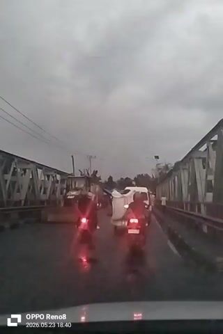 A video taken from inside a car shows traffic on a metal truss bridge on a cloudy day. The road appears wet.  In front of the car, there are several motorcycles and a white van. Further up the road, a yellow Caterpillar construction vehicle with a front-end loader is parked, partially blocking the lane. Pedestrians can be seen on the side of the road. Power lines run alongside the bridge. The video was apparently recorded on an OPPO Reno8 on May 23, 2026, at 18:40.