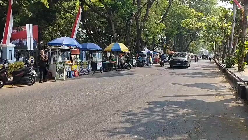 The video shows a street scene with several food stalls set up along the side of the road. A black car is driving down the street, followed by several motorcycles. The stalls have colorful umbrellas and are displaying various items for sale. There are also some people standing around the stalls. The road is paved and lined with trees, and there are flags displayed on poles along the sidewalk. The scene appears to be a busy marketplace or a street festival.