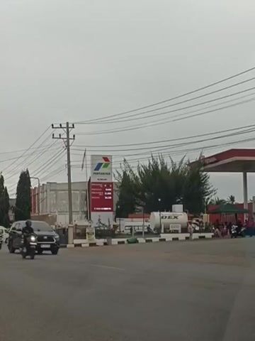 A street-level view shows a Pertamina gas station under a cloudy sky. A black SUV drives past the foreground on the right, while a motorcycle is visible in the middle ground to the left. To the left of the gas station, a white truck drives by. The gas station has a large red and white sign with the Pertamina logo, and several gas pumps are visible. People are gathered around the gas station.