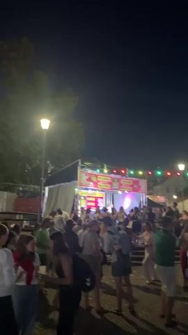 The video shows an outdoor festival at night. A crowd of people are gathered in front of a stage with colorful lights. Some people are dancing, while others are standing and talking. There are string lights strung across the area, and a few tall poles with flags are visible in the background. The atmosphere appears lively and festive.