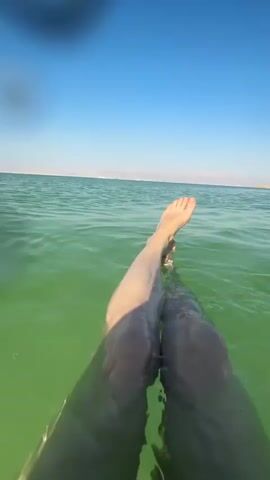The video shows a first-person perspective of someone's legs floating in green water. The camera is submerged in the water, and the legs are extended upwards, with the feet visible above the surface. The legs are fair-skinned, and the toenails are unpainted. The water is clear enough to see the texture of the skin on the legs. The background shows a clear blue sky and a distant shoreline with mountains. There are also some blurry white structures in the background, possibly part of a pier or a beach facility. The overall impression is one of relaxation and buoyancy.