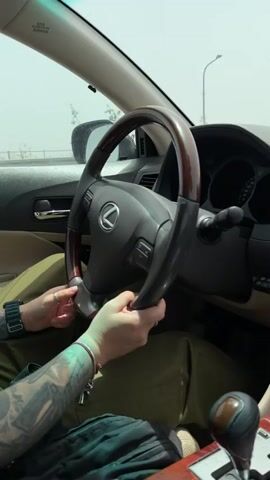 From the driver's seat of a Lexus, an individual drives on a somewhat hazy day. The Lexus features a wood-trimmed steering wheel and beige interior. The driver has visible tattoos on their left forearm, and wears a watch and rings. They have both hands on the steering wheel, guiding the car forward. The outside scenery, seen through the windshield, consists of a highway setting with street lights in the distance. The video presents a first-person perspective from inside the car.