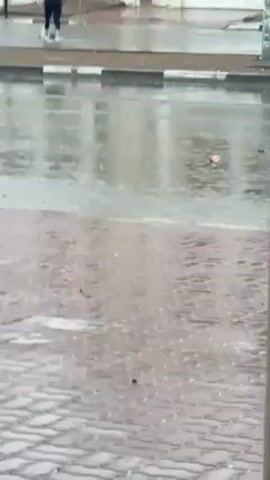The video shows a street scene on a rainy day.  Water covers the road and sidewalks.  Several people are visible, including a man in dark clothing who is initially bent over and then stands up, tossing an object into the air. Another person, who appears to be a younger male, is standing near a carport.  Other people can be seen further away, near buildings.