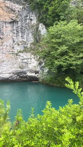 This video shows a serene natural landscape with a vibrant turquoise lake nestled against a towering, rocky cliff face. Lush green foliage blankets the upper portion of the cliff and spills over the edges, creating a dramatic contrast with the pale, textured rock. The water is calm and reflective, with a deep blue-green hue.

In the foreground, a variety of green plants and bushes, some with small white flowers, frame the scene, adding to the sense of natural beauty. On the left side of the lake, a small waterfall trickles down, feeding into the main body of water.

Several people are present near the waterfall, some standing and observing the scenery, while others appear to be engaged in conversation or taking photographs. One person is wearing a white outfit and another is in casual clothing. A large, colorful umbrella provides shade over a seating area. On a small rocky outcrop in the lake, a peacock is visible, adding a touch of exotic wildlife to the picturesque setting. A small palm tree also grows on this outcrop.

The overall impression is one of a peaceful and visually stunning natural attraction, likely a tourist destination given the presence of people and amenities like the umbrella and pathways. The time of day appears to be daytime, with good lighting. The weather seems pleasant, suitable for outdoor activities. The emotions evoked are likely awe and tranquility. No visible text is present in the video.