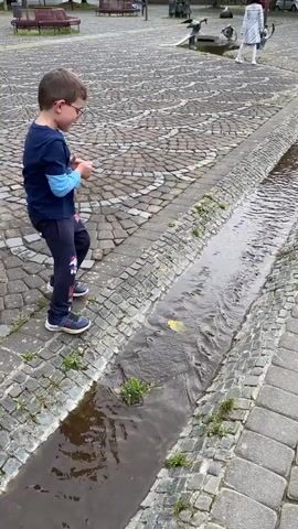 A young boy wearing a blue shirt and dark pants stands on a cobblestone path beside a narrow water channel. A yellow leaf floats in the water. The boy is looking at the leaf. In the background, two adults are visible, one in a light-colored outfit and the other in dark clothing. The environment appears to be an outdoor public space with trees and benches in the distance. The boy is facing away from the camera. The water channel is lined with stone. The cobblestone path has a pattern.