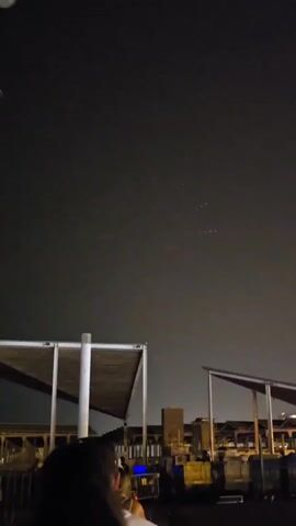 This video captures a night scene from behind a group of people, observing a drone light show. The drones form a large, circular, starburst pattern of white lights against a dark sky. The foreground shows the backs of several people's heads, with a section of a covered structure, possibly a pavilion or seating area, to their left. Beyond the people and the structure, city lights are visible, with some buildings and industrial-looking elements. The drone display is the central focus, with the lights creating a dynamic, illuminated pattern that changes slightly between frames.