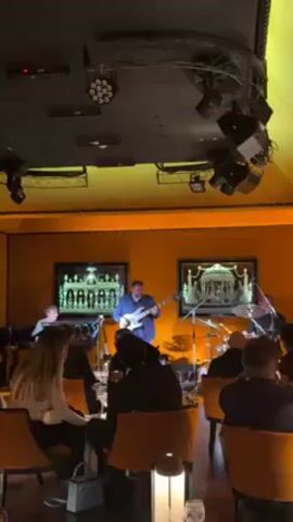 The video shows a live band performing for an audience in a dimly lit restaurant. The band consists of four musicians: a keyboard player, a bassist, a drummer, and another musician who is obscured by the others. The bassist is in the center of the frame, playing his instrument while standing. The audience is seated at tables, enjoying the music. The restaurant has orange-colored walls with framed artwork. The ceiling is dark and features an array of lighting equipment. The camera is static throughout the video.