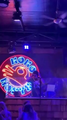 The video shows a DJ mixing music at a venue called Koko. He stands behind a clear partition in front of a neon sign with the venue's name and a graphic of a hand. The lighting is dark with neon highlights in pink, red, and blue. He appears to be wearing headphones and looking at his equipment. A laptop sits on the deck in front of him. A ceiling fan is visible, and some of the crowd can be seen below him.