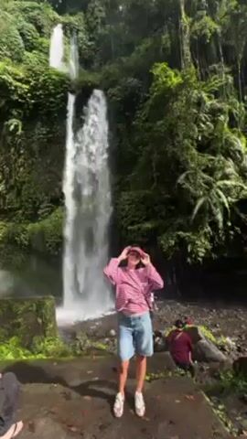 The video captures a woman standing in front of a waterfall. She is wearing a pink and white striped shirt, jean shorts, and sandals, with a pink visor on her head. She is holding her hands up and dancing in front of the three tiered waterfall, surrounded by lush green vegetation. There are other people and objects visible in the scene, but they are not the main focus.