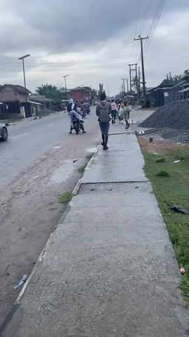 A video shows a street scene on a cloudy day. The camera is positioned on a concrete sidewalk facing down the street. On the left side of the street is a paved road with a silver sedan driving away from the camera and several motorbikes. On the right side of the street, there is a pile of gravel. People are walking on the sidewalk and road. One person, wearing a backpack, walks away from the camera on the sidewalk. Another person, carrying items on their head, walks towards the camera on the sidewalk. Further down the street, more people are visible, along with buildings lining the street. Utility poles with wires are present on the right side of the street.