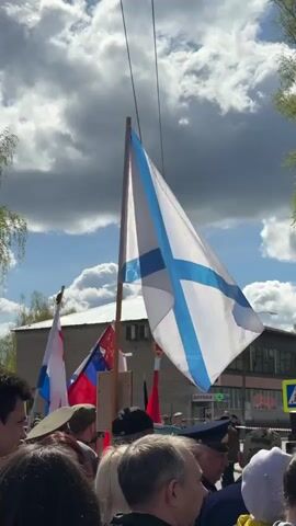 The video shows a public event with a crowd gathered in an outdoor setting. The scene includes a white flag with a blue cross blowing in the wind, a Georgian flag, and Soviet flags. Several people are visible in the crowd, some wearing military-style hats. A building labeled "APTEKA" (pharmacy) is visible in the background, along with trees and a partly cloudy sky. A crosswalk sign indicates a street nearby. People stand in front of a microphone.