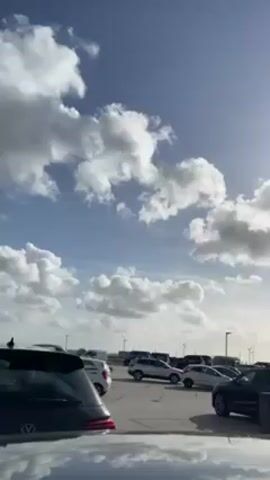 The video shows a parking lot under a bright blue sky with scattered, fluffy white clouds. The sun is shining through the clouds, creating a slight glare. Various cars, mostly dark colors like black and gray, are parked in rows, filling the parking lot. Streetlights are interspersed between the cars. In the distance, there are multiple wind turbines. The camera appears to be positioned low, possibly on the roof of a car, as the reflection of the sky and clouds is visible.