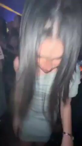 A short video shows a young woman dancing in a crowded, dimly lit nightclub or party venue. Initially, the camera is very close to her face and long, dark hair, obscuring her features. As she dances, she ducks down and then stands up, revealing that she is wearing a short, light-colored dress. Other people, mostly women, are visible around her, also dancing and holding drinks. The lighting is dynamic, with shifting blue and purple hues. The atmosphere seems lively and energetic. The focus is on the woman dancing, with the other individuals appearing as part of the background of the social event.