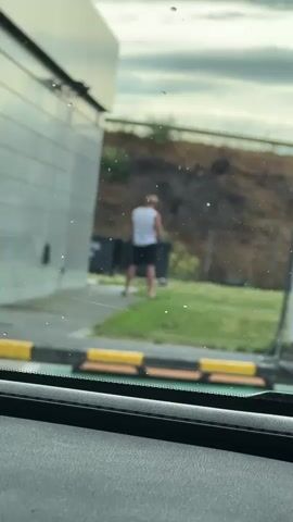 The video captures a person in a straw hat, white tank top, and black shorts standing near trash bins. The person appears to be urinating. The scene is outdoors, with a building to the left and a grassy area with some debris behind the person. The foreground shows the interior of a vehicle, with the dashboard and windshield visible. The lighting suggests it is daytime, with an overcast sky.