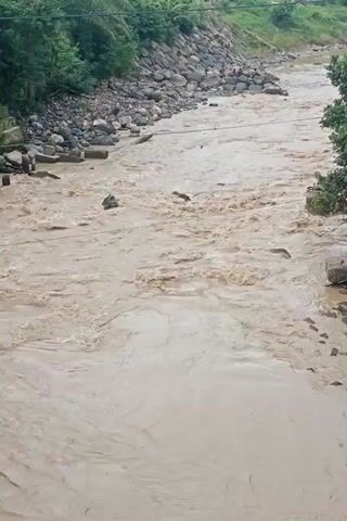 The video shows a river in a state of flood. The water is a muddy brown color, indicative of a high sediment load. The riverbanks are lined with rocks and vegetation, with some trees visible. The water is flowing swiftly, indicating a strong current. The video is taken from a high vantage point, possibly a bridge or elevated embankment.
