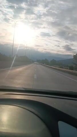 The video shows a first-person perspective from inside a vehicle, looking out at a highway. The sun is low in the sky, casting long shadows and creating lens flare. The sky is partly cloudy. The road appears to be a multi-lane highway with guardrails and streetlights visible on the side. There are trees and some hills or mountains in the distance. There are other vehicles on the highway, but they are far away and not clearly discernible. The interior of the vehicle is dimly visible in the foreground, including part of the dashboard.