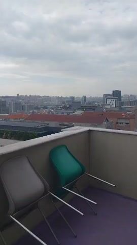 The video presents a view from a rooftop looking out over a city on a cloudy day. Two plastic chairs, one grey and one green, are positioned in the foreground, partially obscured by a low wall. In the background, a dense urban landscape unfolds, composed of buildings with varied architectural styles, primarily featuring red rooftops. A distinct oval-shaped building stands out in the city skyline. The sky is overcast with a uniform layer of grey clouds, lending a muted tone to the scene.