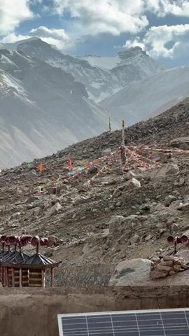 The video shows a mountainous landscape with a rocky hillside adorned with colorful prayer flags. In the foreground, there is a small structure resembling a gazebo with several speakers attached to its roof. A solar panel is also visible, suggesting a remote or off-grid location. The background features towering mountains, partially covered in snow, under a sky with scattered clouds. The overall scene evokes a sense of tranquility and isolation, with the prayer flags adding a touch of cultural and spiritual significance.