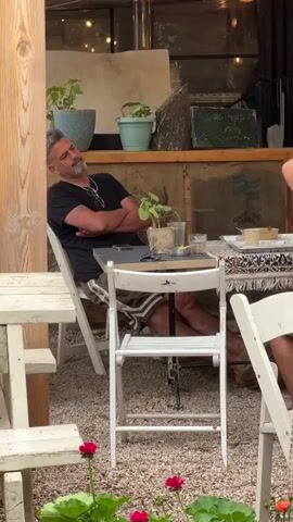 Here's a description of the video:

The camera pans across an outdoor cafe area.  A man with graying hair and a beard is sitting at a table, leaning back in his chair with his arms crossed.  Another, younger man sits down at a nearby picnic table.  There are two ice cream cones upside down on the table.  The tables are white wood and there are potted plants and flowers scattered around. A closed beige umbrella is visible.  The ground is covered in small pebbles.