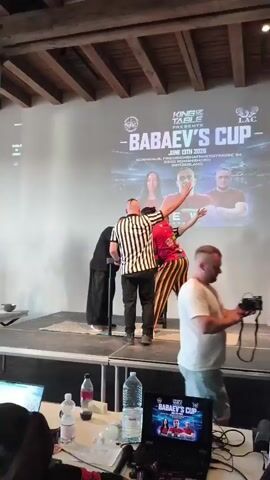 The video opens on an arm wrestling match stage. A referee in a black and white striped shirt stands between two arm wrestlers, both of whom are wearing distinctive clothing. The wrestler on the left, wearing a black outfit, is gripping the arm wrestling device. The wrestler on the right, wearing a bright red t-shirt with cartoon characters and black and yellow striped pants, is in position to compete. Behind them, a large screen displays "KING OF TABLE PRESENTS - BABAEV'S CUP JUNE 13TH 2026 KORNHAUS, FRIEDRICHSHAFNERSTRASSE 54 8590 ROMANS HORN SWITZERLAND". The referee signals to the competitors. The wrestler in red raises his arm triumphantly, then walks away from the table. The other referee, who was previously obscured, is now visible. The wrestler in red continues to walk off stage, acknowledging the crowd. The screen behind them shows images of two men and a woman. The arm wrestling table is positioned on a raised platform.