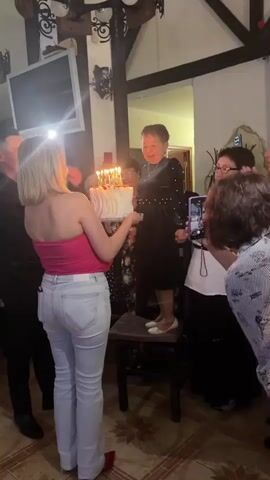 A group of people are gathered around an older woman who is standing on a chair, holding a birthday cake with lit candles. A younger woman in a pink top and light-colored jeans is holding the cake for the older woman. Several people are taking photos and videos with their phones. The setting appears to be an indoor gathering, possibly a restaurant or home, with decorative elements on the ceiling.