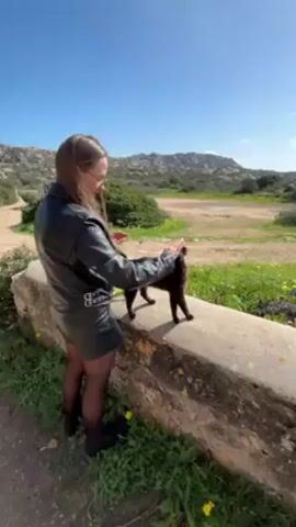 A woman is seen petting a black cat in an outdoor setting. The woman, wearing a black leather jacket and matching skirt with dark tights and boots, is standing next to a low, stone wall. She is bent slightly, her hands petting the cat's back. The cat is standing on the wall, arching its back into the woman's touch. The background reveals a sunny landscape with a grassy field, sparse vegetation, and rocky hills under a clear, blue sky. A dirt path can be seen running along the left side of the scene.