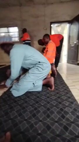 Here is a description of the video:

The video shows four people in a room that appears to be a prayer space. Three individuals are actively engaged in prayer, kneeling and bowing on a patterned rug. The person in the foreground is dressed in a light blue garment, while another wears yellow clothing. Two other individuals are wearing bright orange safety vests. One of the men in the safety vests is kneeling and seated on his legs in prayer, while the other is standing. A small child is next to the kneeling man. A window with a visible outside environment is behind the standing man. The room appears modestly decorated, with light walls. A doorway is visible towards the right side of the frame.