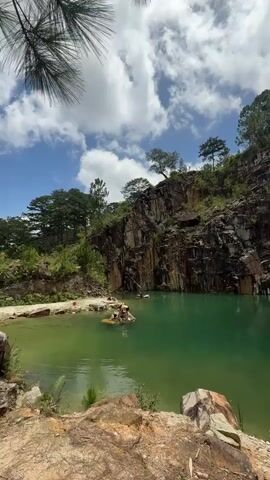 The video shows a serene lake with turquoise water surrounded by a rocky cliff face. Lush green trees grow on top of the cliff and around the edges of the lake. The sky is a bright blue with scattered white clouds. In the lake, a small group of people are enjoying themselves on an inflatable raft. The scene appears to be a natural, secluded spot, perhaps a quarry or a natural basin, offering a peaceful and picturesque escape.