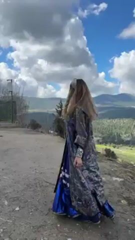 A woman in a long, patterned blue and gray coat and blue dress spins around. She has long blonde hair and is wearing a white top under the coat and dress, along with jewelry. She is standing on a dirt surface overlooking a green valley with trees and mountains in the background. The sky is blue with white clouds.