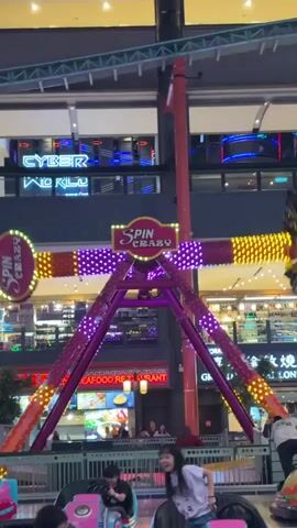 The video shows an indoor amusement park ride called "Spin Crazy" in action. The ride features a central vertical support with a rotating arm extending horizontally, holding passenger cars on either end. The arm swings in a circular motion, lifting the cars high in the air. The ride is adorned with brightly colored lights, including yellow, red, and purple. Below the ride, a metal safety fence surrounds a children's ride that appears to have no current riders. Several people are visible in the background, including those waiting for or watching the ride. Above, there is a neon sign that says "Cyber World". The environment appears to be a mall or shopping center, with shops and restaurants visible in the background.