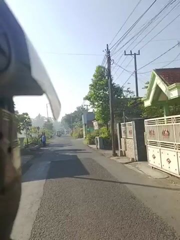 A first-person view from a moving vehicle, likely a motorcycle, shows a narrow asphalt road in a rural or semi-urban setting under a clear, bright sky. On the left side, a person in a light blue top stands near the roadside with trees, plants, and a blue barrel. A street light pole is also visible on the left. On the right side, houses and buildings with various potted plants and dense foliage line the road. Overhead power lines span across the road, connected to utility poles on the right. Further down the road, other vehicles, including a larger truck, are visible in the distance.