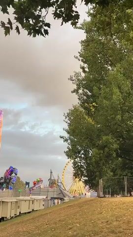 The video shows a Ferris wheel at a fairground against a cloudy sky. A large tree with green leaves is on the right side of the frame, partially obscuring the Ferris wheel. There are other carnival rides visible in the background, including what appears to be a drop tower or roller coaster. The Ferris wheel is lit up with yellow and green lights. In the foreground, there is a grassy slope and a white fence. Several white portable buildings are lined up on the left side of the frame. The overall scene depicts a lively amusement park setting.