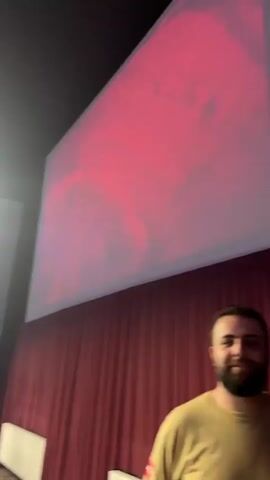 A man with a beard smiles at the camera. He is standing in front of a large screen that displays an image of green clouds and the red letters "I'M". A red curtain hangs behind the screen. The scene appears to be indoors, possibly in a theater or auditorium.