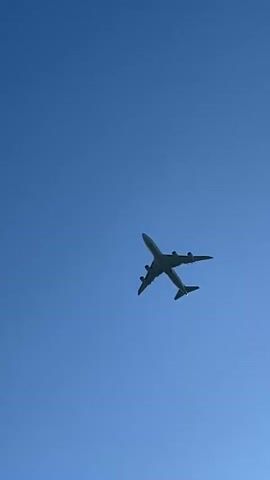 The video captures a large, four-engine passenger airplane flying high overhead against a clear, brilliant blue sky. The aircraft is viewed from below, appearing to ascend or maintain a high altitude. Its silhouette is visible against the bright sky, and the four engines are discernible under its wings. There are no clouds, other objects, or visible text in the sky or on the plane. The scene takes place during the daytime in Reckange-sur-Mess, Luxembourg.
