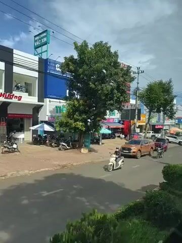 This video shows a street scene in a town or city. There are several buildings on the left side of the street, some of which appear to be businesses. There are trees along the sidewalk and a street with cars and motorcycles. In the foreground, there are some bushes. The sky is cloudy. On the right side of the street, there is a lamppost with a banner attached to it. A white electric car is driving down the street.