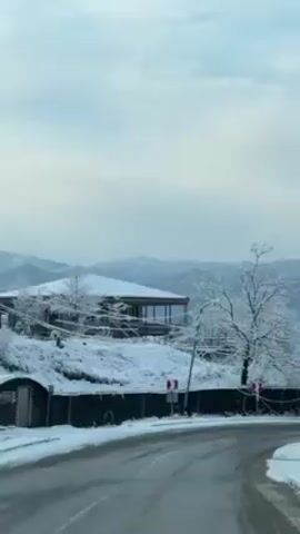 The video shows a view from inside a vehicle traveling on a road in a snowy, mountainous landscape. The road is wet and partially clear of snow, while the surrounding area is covered in a thick layer of snow. There is a building on a snowy hill to the left of the road and bare trees covered in snow throughout the landscape. A silver or white car is parked off the side of the road, almost hidden among the snow-covered trees and a fence. Power lines and poles run along the side of the road. The sky is overcast with a light blue hue. The overall scene is serene and depicts a winter landscape.
