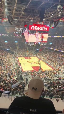 A basketball game is being played at an indoor arena. The stands are filled with spectators. The Cleveland Cavaliers are playing the Houston Rockets. The Cavaliers are in the lead 30 to 16 with 3:39 remaining in the first quarter. There is a man sitting in the stands facing the court wearing a white baseball cap and a black t-shirt that reads "D & S Heating & Cooling". He is holding a plate with food. A jumbotron hangs above the court. The game action is visible on the jumbotron.