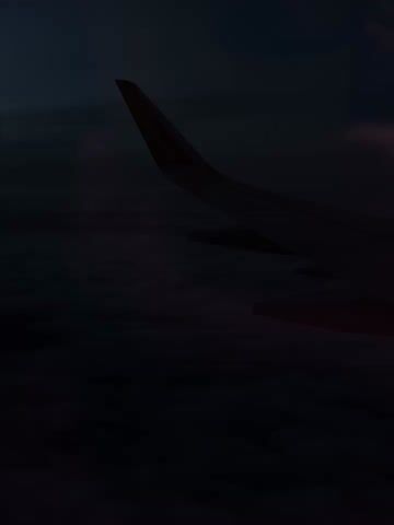 The video captures a view from an airplane window, looking out over a vast expanse of clouds. The dark silhouette of an airplane wing dominates the right side of the frame, gradually becoming clearer as the video brightens. The wing features a red winglet with the text "wizzair.com" visible on its surface, indicating a Wizz Air flight. Below the wing, dense, dark grey-blue clouds stretch out to the horizon, suggesting the aircraft is flying above a cloud layer. The sky above is a deep, muted blue. Pink and purple light flares or reflections are scattered across the frame, particularly noticeable in the upper left and lower right, likely from internal cabin lights reflecting off the window. The overall impression is that of a flight during twilight or nighttime, with artificial light contributing to the atmospheric glow.