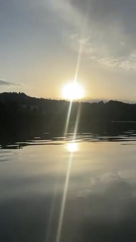 The video opens with a shot of the sun setting over a body of water. The sun is bright and appears to be low in the sky, casting a golden glow on the water and the surrounding landscape. The camera then pans to show a person sitting in a pedal boat, their legs visible in the foreground. The person is wearing ripped jeans. The boat is on a lake, and in the background, there are trees and buildings along the shore. The sun continues to set, and the sky is filled with soft clouds. As the video progresses, the boat moves closer to the shore, revealing more details of the buildings and the surrounding area. There are several buildings visible, including what appears to be a large, ornate castle-like structure. The water is calm, reflecting the sky and the surrounding scenery. The overall atmosphere is peaceful and serene.