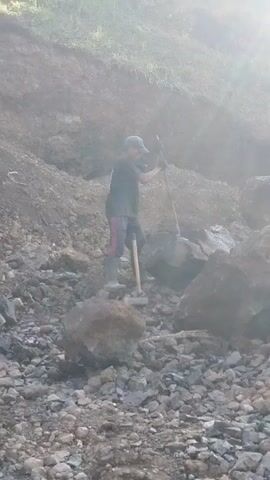 A person is shoveling rocks on a hillside. The person is wearing a black t-shirt, pink pants, black boots, and a cap. The person is using a shovel to move rocks. The hillside is covered in rocks and dirt. The sun is shining brightly, creating a lens flare effect.