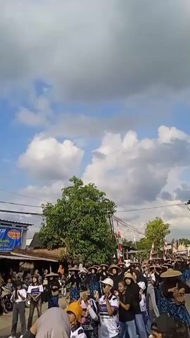 A group of people wearing similar clothing and hats are walking down a street. Some onlookers are standing along the side of the street watching them. There are buildings on the side of the road and a tree in the middle. The sky is blue with scattered clouds. A sign for "Armaham Plastic Shop" is visible. There are Indonesian flags displayed. People are taking pictures with their phones.
