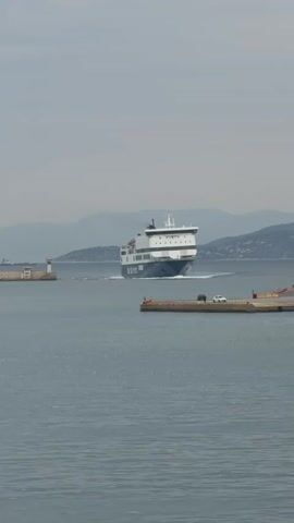 The video shows a large ferry traveling through a body of water on a cloudy day. The ferry is white and blue with the name "Blue Star" visible on its side. It is moving towards the viewer, creating a wake behind it. In the background, there are distant mountains. To the left of the ferry is a small lighthouse, and to the right, there is a dock with two cars parked on it.