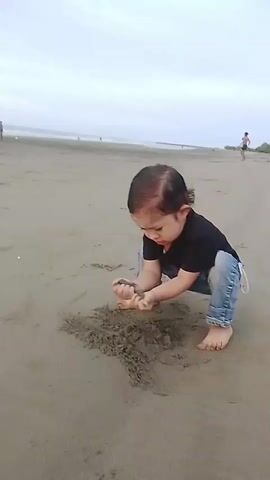 A young child wearing a black t-shirt and blue jeans is squatting on a sandy beach and playing with the sand. The child is holding some sand in their hands and appears to be drawing in the sand with their fingers. In the background, there is the ocean and other people on the beach. The sky is overcast.