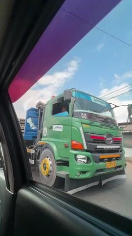 The video shows a green HINO truck with the text SARANA on the side, stopped on a road. The driver, a person wearing a blue shirt, is visible inside the cab. The truck has a blue container on its back. The image is taken from inside another vehicle, with part of the window frame visible in the foreground. The sky is blue with some clouds. Electrical wires are visible in the background.