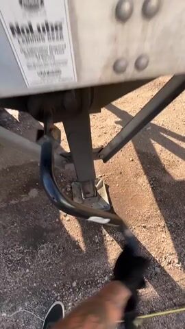 The video shows a close-up of a semi-trailer landing gear, with a person's feet visible at the bottom of the frame. The landing gear is a heavy-duty metal jack used to support the trailer when it's unhitched. The ground is covered in gravel and dirt, suggesting an outdoor, possibly industrial or truck yard setting. Sunlight casts long shadows, indicating it's likely late afternoon or early morning.

A label on the landing gear reads "NoLube LANDING GEAR" and appears to have a brand name above it. Another label, partially visible on the left, has text that begins with "ATTENTION" and provides some warnings or instructions. The trailer itself is white with red reflective tape visible at the top edge.

The person's feet, wearing black sneakers, are positioned near a yellow object on the ground, possibly a measuring tape or boundary marker. The overall impression is one of preparation or inspection related to a truck or trailer. No specific emotions are conveyed, and the activity seems to be static, focusing on the equipment. The location context provided is Aurora, United States of America.