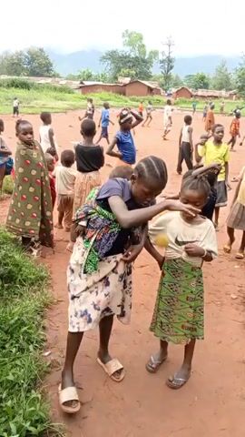 A daytime, eye-level, medium-shot video shows a group of children on a dirt field.  In the foreground, a young woman wearing a patterned skirt, a navy shirt, and sandals is carrying a small child on her back wrapped in a colorful cloth. To the right of her, a young girl in a white t-shirt and green patterned skirt stands with her hands on her hips.  In the background, many other children are playing and moving around on a dirt field. Some are running, while others are standing and observing.  The field is bordered by green grass and foliage.  Beyond the field, there are a few simple buildings and trees under a cloudy sky.